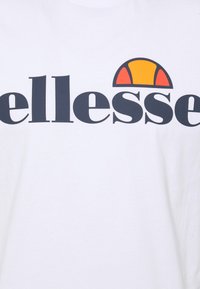 White t-shirt with large black text spelling "ellesse" and a colorful logo featuring a semi-circle in orange and yellow. Soft cotton texture.