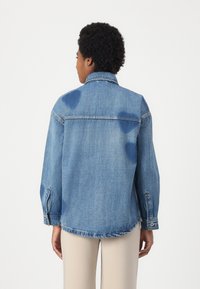 Lyseblå denimjakke med avslappet passform, knappe mansjetter og klassisk krage. Har falmede områder og en buet kant.