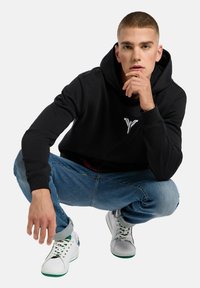 Schwarze, gekürzte Kapuzenjacke mit einem grafischen Logo auf der Vorderseite, kombiniert mit hellblauen Jeans und weißen Sneakers mit grünen Akzenten. Glatte Stoffoberfläche.