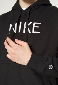Osoba ubrana w czarną bluzę Nike z kapturem, trzymająca jeden czarny sznurek przy białym logo na piersi, mały biały haftowany symbol na rękawie.