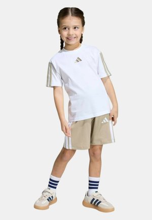 Ragazza giovane con i capelli intrecciati che indossa una maglietta bianca Adidas, pantaloncini beige Adidas, calzini a righe e scarpe da ginnastica, in piedi e sorridente.