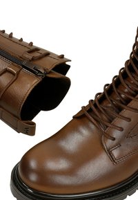 Stivali ankle in pelle marrone con lacci distintivi, zip sul lato e superficie testurizzata. Presentano una punta arrotondata e una suola robusta.