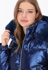 Lesklá námořnická puffer bunda s umělou kožešinou na kapuci, s předním zipem a prošívanou strukturou. Modelka drží límec.