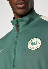 Zelena sportska jakna na zip s okruglim logotipom VfL Wolfsburga, mekani materijal, kontrastni svijetli naglasci i dizajn s visokim ovratnikom.
