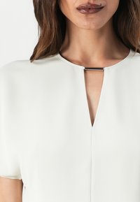 Witte blouse met een keyhole-halslijn en een zilveren horizontale accent. Zachte, gladde stof met korte mouwen en een losse pasvorm.