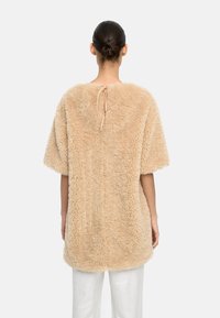 Beżowy top z futra sztucznego, o luźnym kroju, krótkich rękawach i wiązaniu z tyłu. Miękka, teksturowana powierzchnia o puszystym wyglądzie.