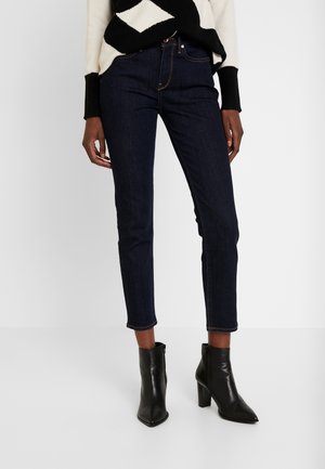 Vrouwelijk model draagt donkere slim-fit enkellange spijkerbroek, zwarte enkellaarsjes met hak en een zwart-wit gebreide trui met patroon.