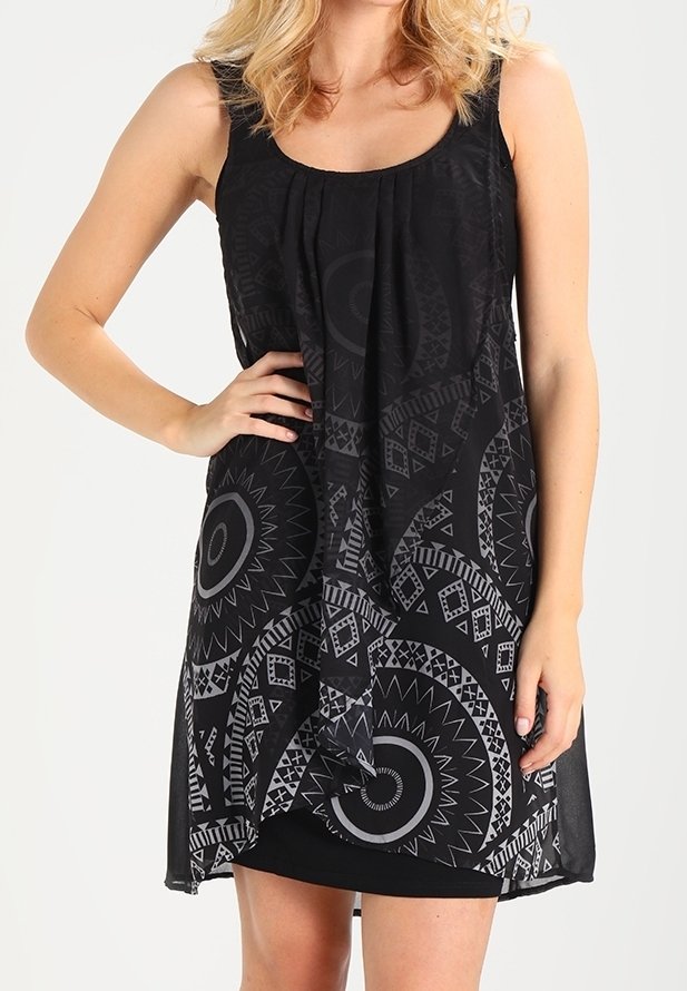 Femme portant une robe noire sans manches avec des motifs géométriques et circulaires gris, une main sur la hanche, debout devant un fond uni.