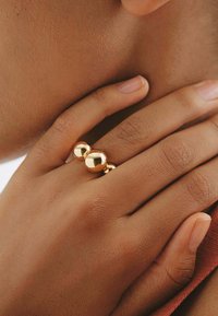 Anillo de oro con tres elementos esféricos pulidos, colocado en una mano con un acabado de uñas natural sobre un fondo de piel texturizada.