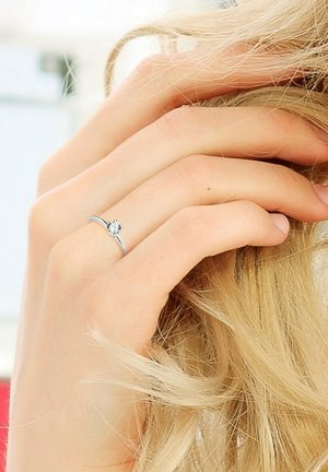 Bague en argent avec un sertissage en pierre précieuse ronde et transparente. L'anneau est fin et poli, reposant sur une main à la peau claire et aux longs cheveux.