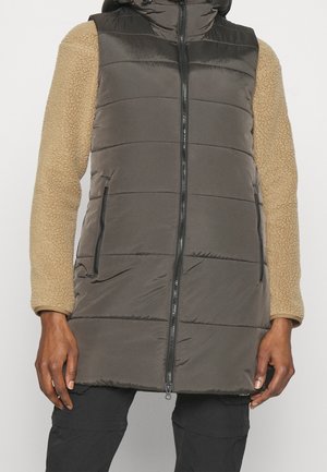 Personne portant une veste matelassée longueur genoux avec fermeture éclair à l'avant, de couleur noire, et des manches longues texturées beige sur fond uni.