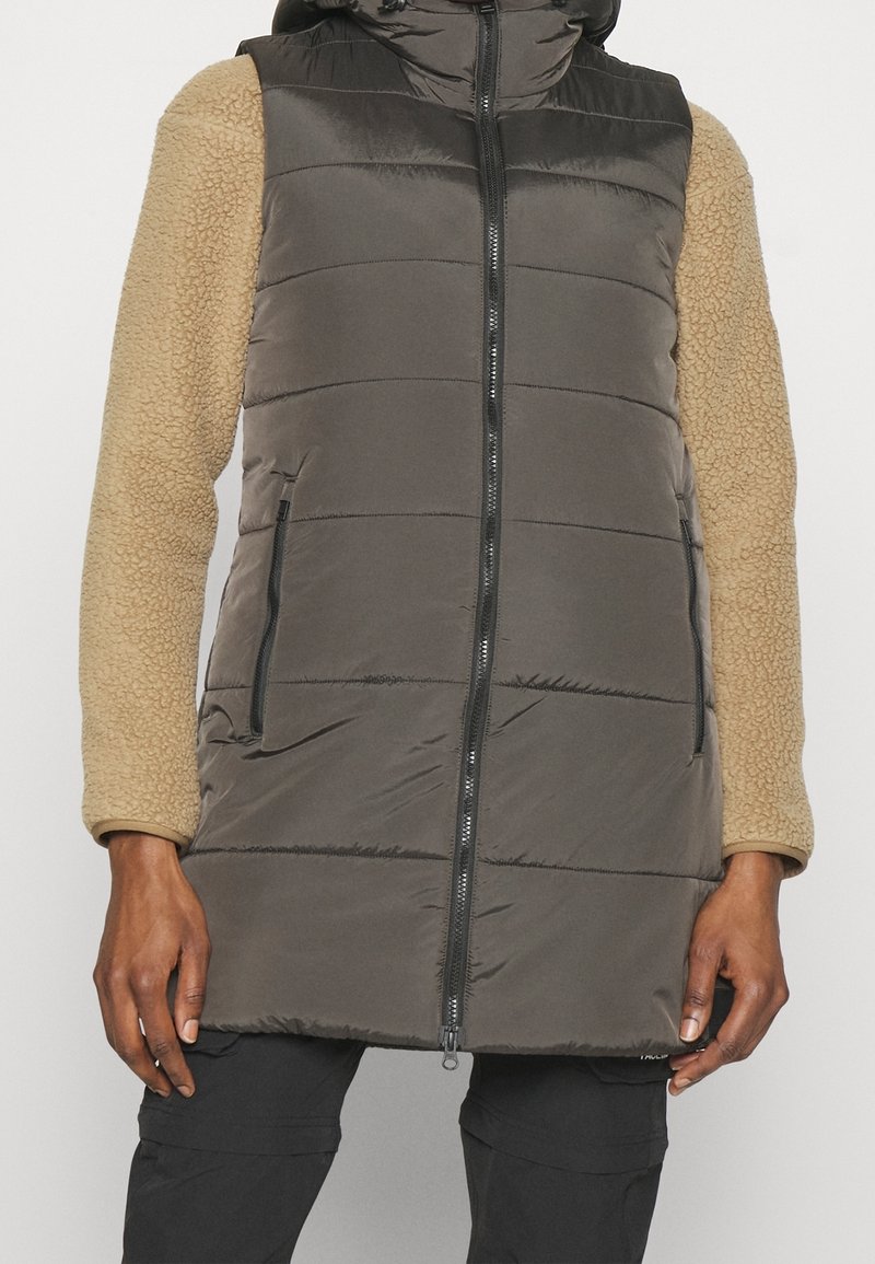 Personne portant une veste matelassée longueur genoux avec fermeture éclair à l'avant, de couleur noire, et des manches longues texturées beige sur fond uni.
