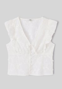 Blouse blanche sans manches avec broderie florale, col en V, épaules à volants, lien devant et fermeture boutonnée sur fond gris.