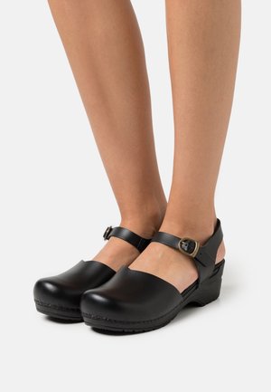 Sabots en cuir noir avec un bout fermé et un talon ouvert. Bride avec une boucle en accent. Talon léger avec base cousue et texture lisse.