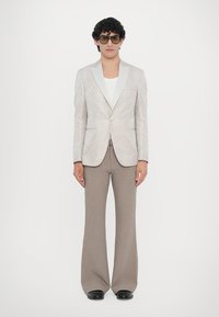 Lightly patterned beige blazer with a satin lapel, paired with light brown flared trousers. Model wears a white shirt and oversized sunglasses.
