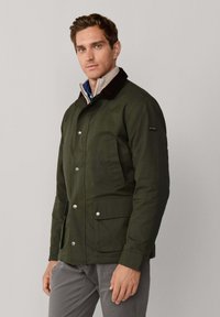 Chaqueta verde oliva con cuello de pana, con botones a presión, dos bolsillos frontales y un forro interior acolchado.