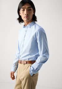 Chemise bleu clair à manches longues avec un col boutonné, associée à un pantalon khaki et une ceinture marron. Présente un logo discret sur la poitrine.
