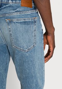 Vaqueros de mezclilla azul claro con textura desgastada, que presentan un bolsillo trasero y una etiqueta de cuero marrón de la marca cerca de la cintura.