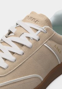 Μπεζ σουέτ αθλητικό παπούτσι με λευκά κορδόνια, διάτρητη πλαϊνή επιφάνεια, καουτσούκ σόλα και λογότυπο ARKK στη γλώσσα και στην πλευρά.