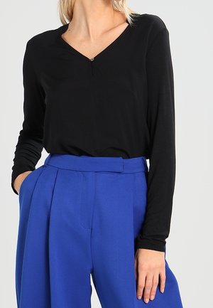 Kobieta w czarnej bluzce z długim rękawem i dekoltem w kształcie litery V, włożonej do spodni o wysokim stanie, plisowanych, jasnoniebieskich, z jedną ręką w kieszeni.