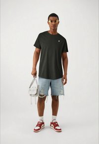 Dark green t-shirt, casual fit. Light blue distressed denim shorts. Red and white sneakers. Clear sunglasses. Gray bag with strap.