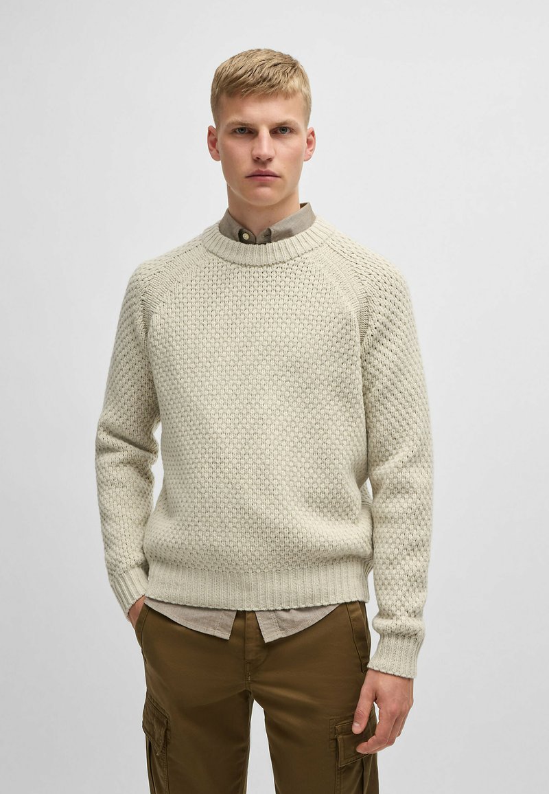 Maglione lavorato a maglia color crema con un motivo testurizzato, con polsini e orlo a coste, indossato sopra una camicia colletto marrone chiaro.