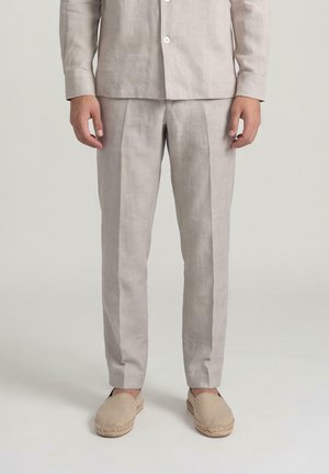 Persoon die lichtbeige linnen broek, bijpassend overhemd met knopen en beige slip-on espadrilles draagt, staand tegen een effen achtergrond.