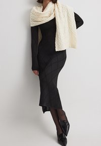 Femme portant une robe en tricot noir texturé, des collants noirs, des mocassins noirs à bout pointu et une grande écharpe en tricot blanc à cable drapée sur les épaules.