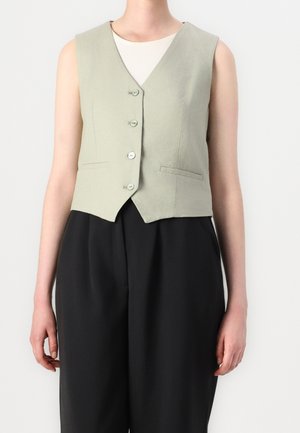 Osoba ubrana w jasnozieloną, bezrękawnikową kamizelkę na guziki na białej bluzce, w połączeniu z luźnymi, czarnymi spodniami z wysokim stanem na jednolitym tle.