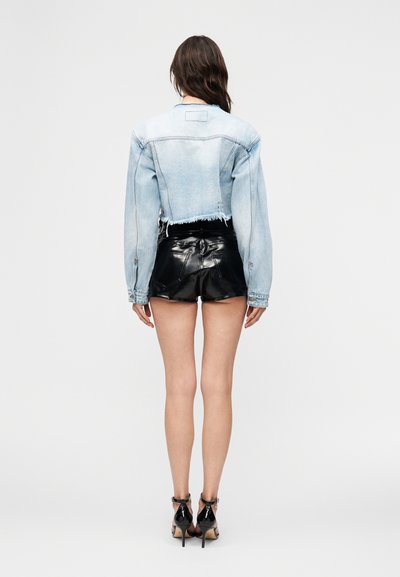 Γυναίκα που στέκεται πλάτη, φοράει ένα ανοιχτό μπλε κοντό τζιν μπουφάν, μαύρα γυαλιστερά δερμάτινα σορτς και μαύρα πέδιλα με ψηλό τακούνι.
