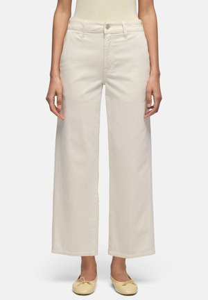 Kobieta w kremowych spodniach z wysokim stanem i szerokimi nogawkami, zapinanych na guzik i suwak, w zestawieniu z beżowymi balerinami na jednolitym tle.