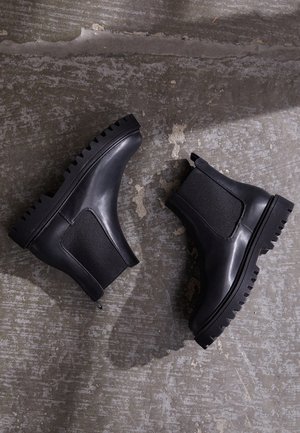 Bottines Chelsea noires avec une tige en cuir lisse, des panneaux latéraux élastiques et une semelle en caoutchouc épaisse avec des rainures profondes pour une meilleure adhérence.