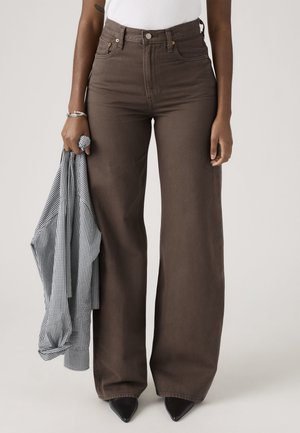 Osoba nosiaca hnedé široké nohavice s vysokým pásom, v ľavej ruke drží čiernobielu kockovanú košeľu, stojí v čiernych špicatých topánkach.