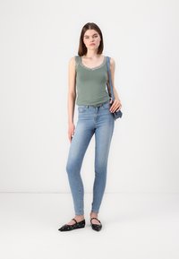 Green tank top with lace trim, paired with light blue skinny jeans. Model wears black flats with decorative studs.