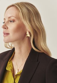 Guldmetalløreringe med en abstrakt form, båret sammen med en grøn bluse og en mørk blazer; glat tekstur med en reflekterende overflade.