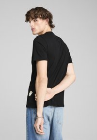 Sort bomulds T-shirt med rund hals, korte ærmer og en etikette i siden. Modellen poserer med hænderne placeret bagpå, iført lyseblå jeans.