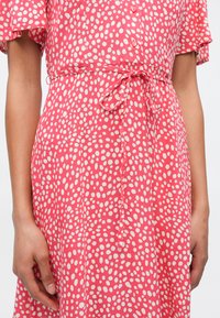 Γυναίκα που φοράει ροζ φόρεμα με λευκές ακανόνιστες πουά, ζώνη στη μέση δεμένη σε φιόγκο και κοντά μανίκια με κυματιστό σχέδιο, στέκεται με τα χέρια χαλαρά στο πλάι.