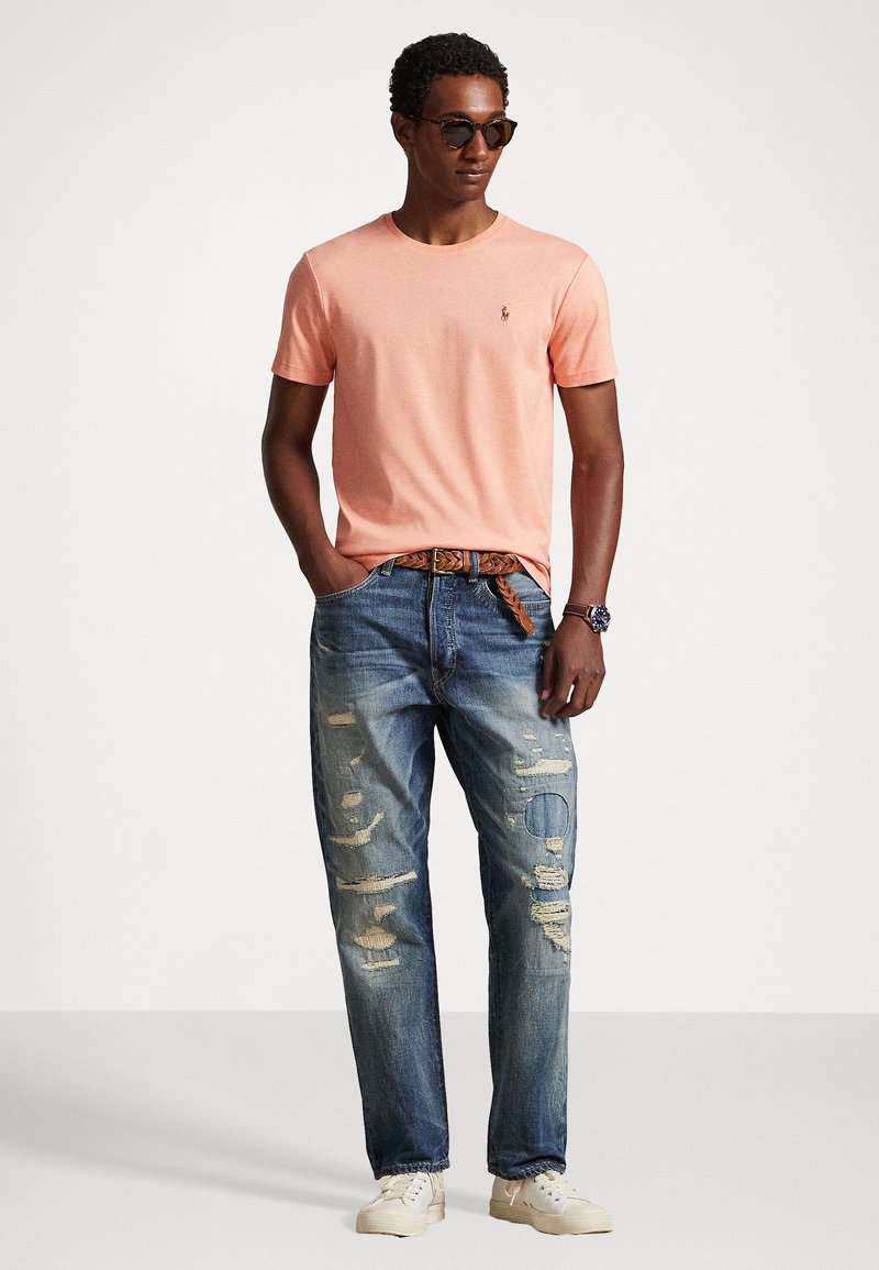 T-shirt corail clair avec col rond, assorti à un jean bleu usé avec un patch circulaire et un léger effet vieilli. Des baskets beiges complètent le look.