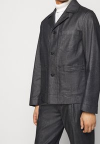 Chaqueta de mezclilla oscura con cuello con muesca, tres bolsillos frontales y cierre de botones. Textura suave con detalles de costura visibles.
