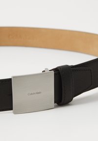 Ceinture en cuir noir à texture lisse, boucle rectangulaire en argent avec logo "Calvin Klein" embossé, et dessous beige.