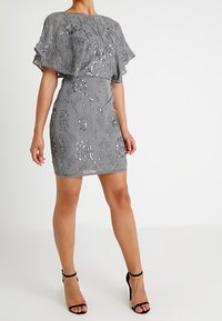 Kobieta w dopasowanej szarej sukience wyszywanej cekinami, z luźnym narzucanym płaszczem i czarnymi sandałami na obcasie z paskiem w okolicy kostki.