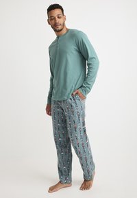 Camiseta de pijama de manga larga en color teal con cuello redondo; pantalones de pijama con patrón de dibujos de cascanueces sobre un fondo azul claro.