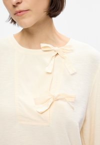 Top de couleur crème avec un tricot texturé, des patches en tissu lisse et deux nœuds décoratifs à l'avant.