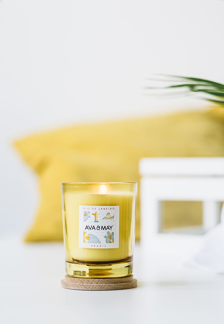 Gelbe Wachskerze in einem klaren Glasbehälter mit einem Holzsockel. Verfügt über ein buntes Etikett, das "Rio de Janeiro" und "Ava & May" beschreibt.