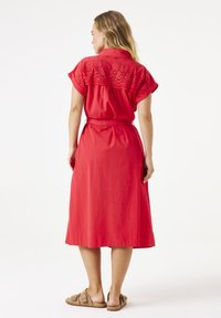 Rode jurk met korte mouwen en borduursel op de rug, strak om de taille, gemaakt van lichte stof. Gedragen met lichtbruine sandalen.