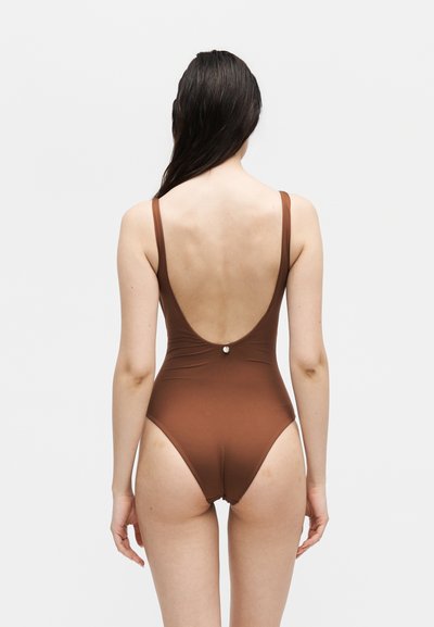 Γυναίκα με μακριά σκούρα μαλλιά που φοράει καφέ ολόσωμο μαγιό με χαμηλή στρογγυλή πλάτη και μια μικρή στρογγυλή μεταλλική λεπτομέρεια.