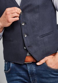 Marinblå herringboneväst med fem knappar, subtil textur, två framfickor, bärs över en mönstrad skjorta, kombinerad med blå jeans och ett brunt bälte.