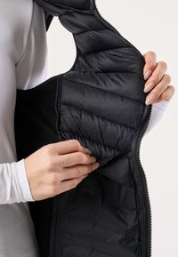 Ruky držia otvorenú čiernu prešívanú bundu, ukazujúc hladký vnútorný materiál a vnútorný vrecko, nosenú cez bielu blúzku s dlhými rukávmi.