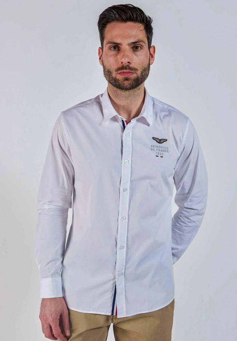 Homme avec une barbe portant une chemise blanche à manches longues boutonée avec le logo "Patrouille de France 1953" et un pantalon beige, standing against a plain background.