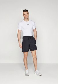 Camiseta atlética branca justa e calções de banho pretos com três riscas brancas. Tênis cinzentos. Design simples e moderno com detalhes de logotipo.